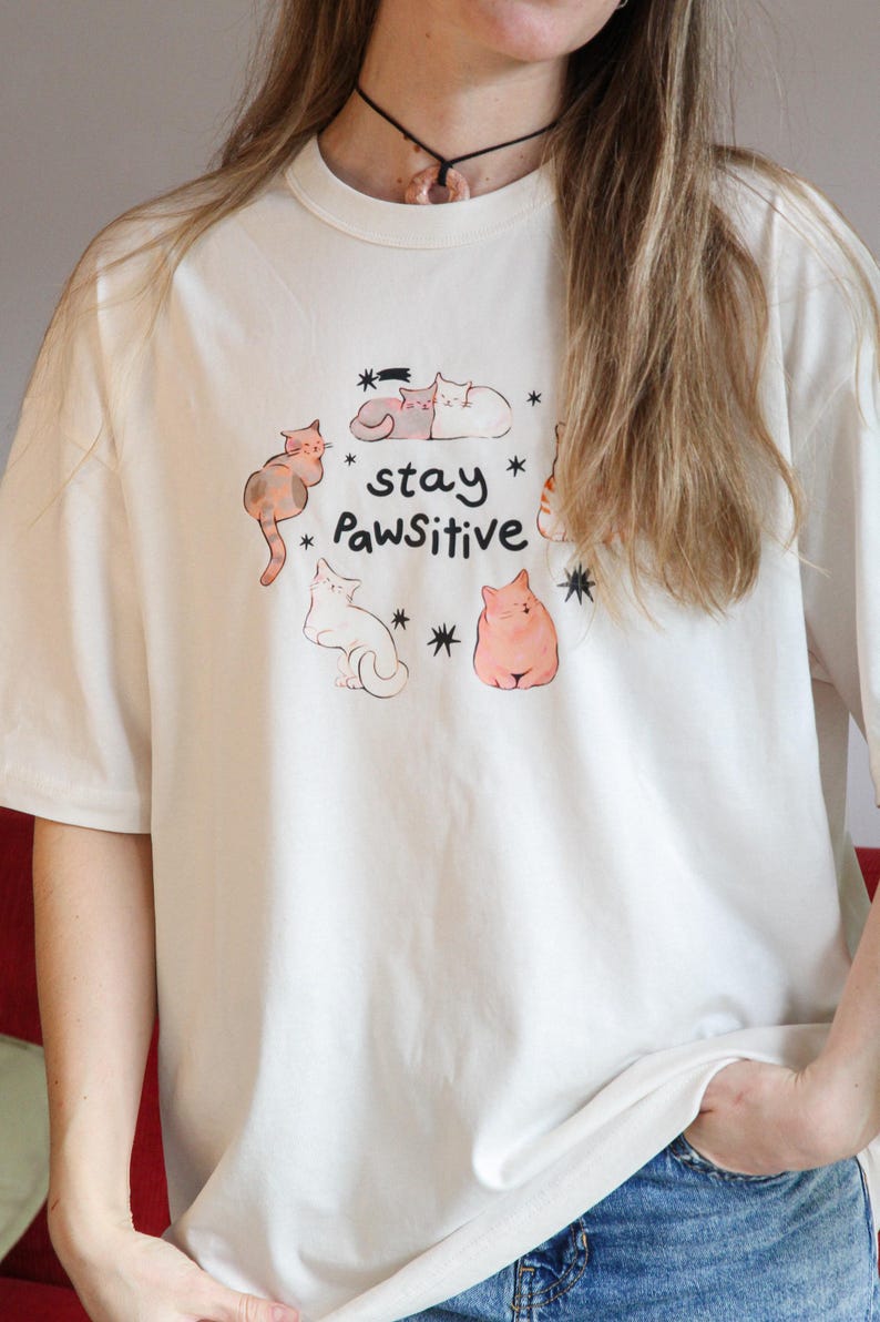 Puede incluir: Camiseta de color crema con un dise&ntilde;o l&uacute;dico de gatos de dibujos animados en varias poses. La camiseta incluye el texto "stay Pawsitive" con acentos de estrellas. Una parte superior informal y c&oacute;moda.