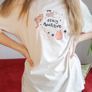 Puede incluir: Camiseta color crema con una ilustraci&oacute;n juguetona de gatos y el texto "stay Pawsitive". El dise&ntilde;o incluye varios dibujos de gatos y estrellas. La camiseta est&aacute; hecha de un material suave y c&oacute;modo.