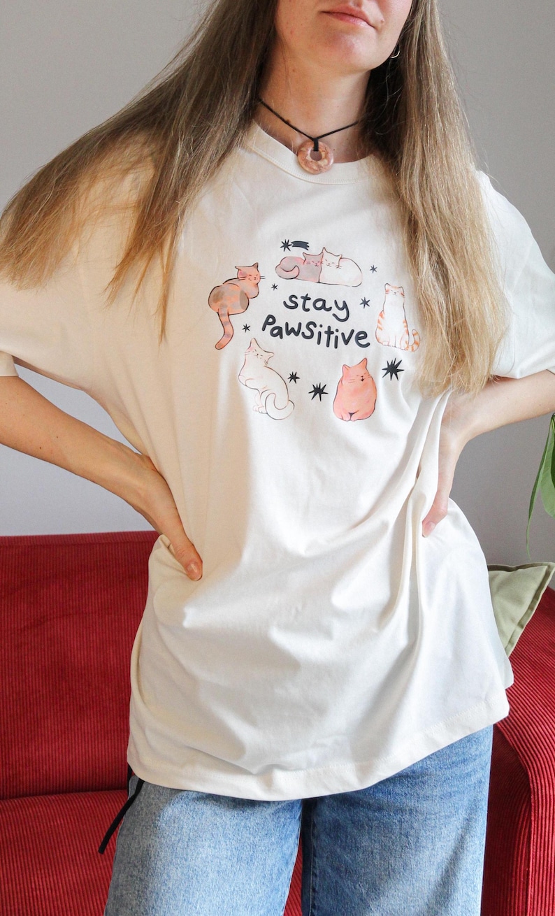 Puede incluir: Camiseta color crema con una ilustraci&oacute;n juguetona de gatos y el texto "stay Pawsitive". El dise&ntilde;o incluye varios dibujos de gatos y estrellas. La camiseta est&aacute; hecha de un material suave y c&oacute;modo.