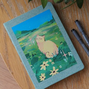Puede incluir: Un diario turquesa con una ilustraci&oacute;n de gato en la portada. La obra de arte representa un gato color crema en un campo de flores bajo un cielo azul. Dos bol&iacute;grafos negros est&aacute;n junto al diario sobre una superficie de madera.