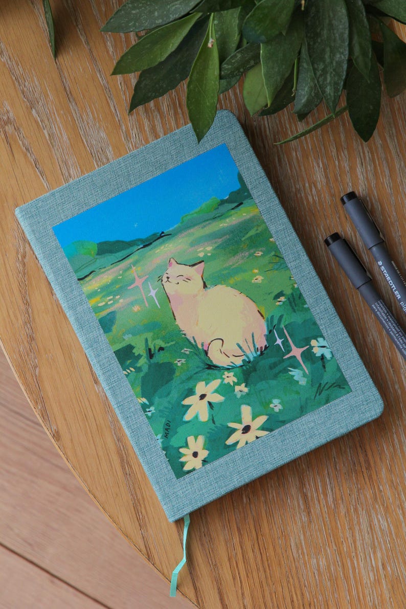 Puede incluir: Un diario turquesa con una ilustraci&oacute;n de gato en la portada. La obra de arte representa un gato color crema en un campo de flores bajo un cielo azul. Dos bol&iacute;grafos negros est&aacute;n junto al diario sobre una superficie de madera.
