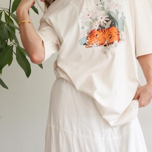 May include: Cream-colored t-shirt featuring a graphic of a sleeping orange tiger surrounded by white flowers and green foliage. The shirt is paired with a white skirt. The overall aesthetic is casual and comfortable.