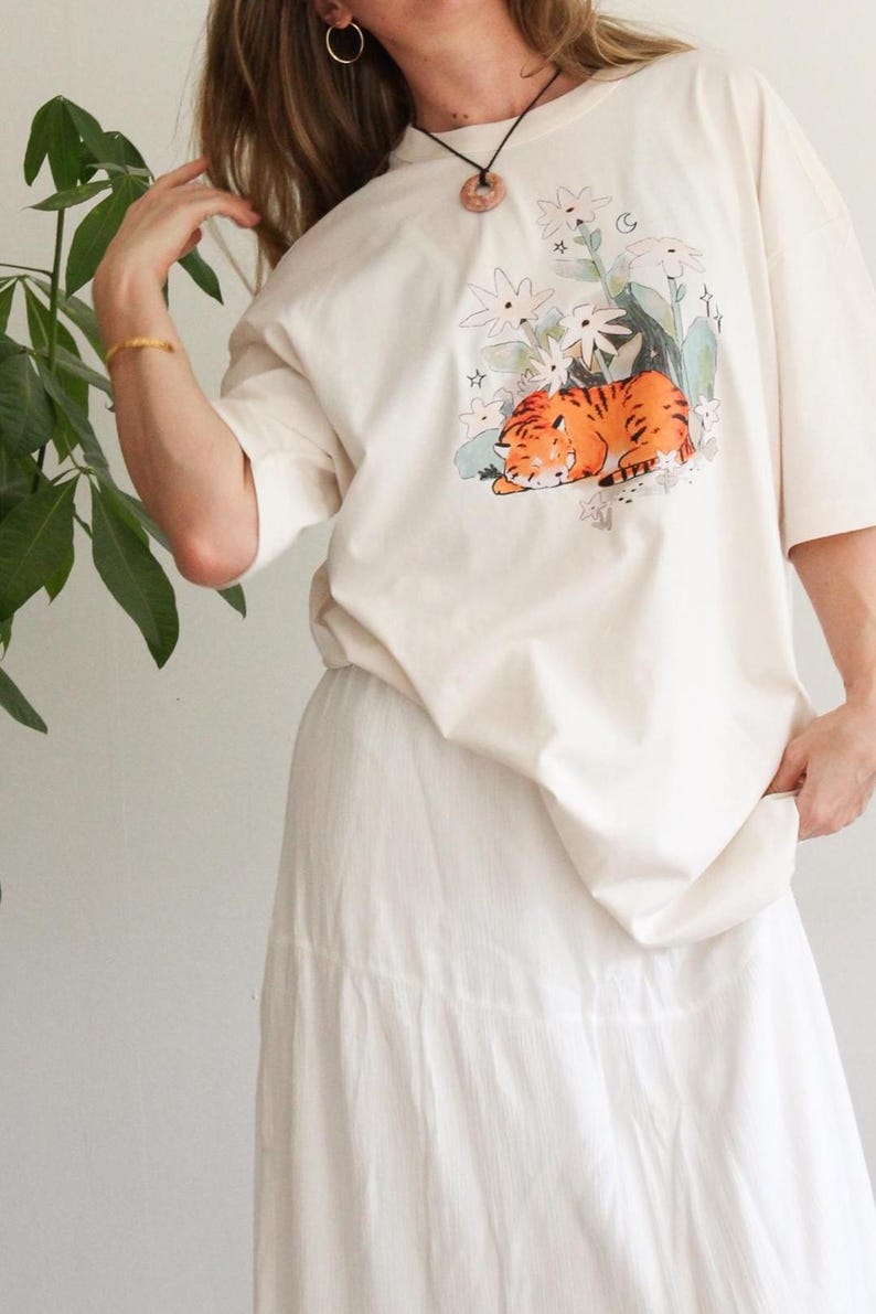May include: Cream-colored t-shirt featuring a graphic of a sleeping orange tiger surrounded by white flowers and green foliage. The shirt is paired with a white skirt. The overall aesthetic is casual and comfortable.