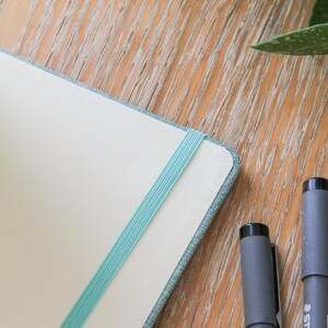 Puede incluir: Un cuaderno con una banda el&aacute;stica azul claro y una cubierta azul claro est&aacute; abierto sobre una superficie de madera. Dos rotuladores grises Staedtler est&aacute;n junto al cuaderno. Hojas verdes en el fondo.
