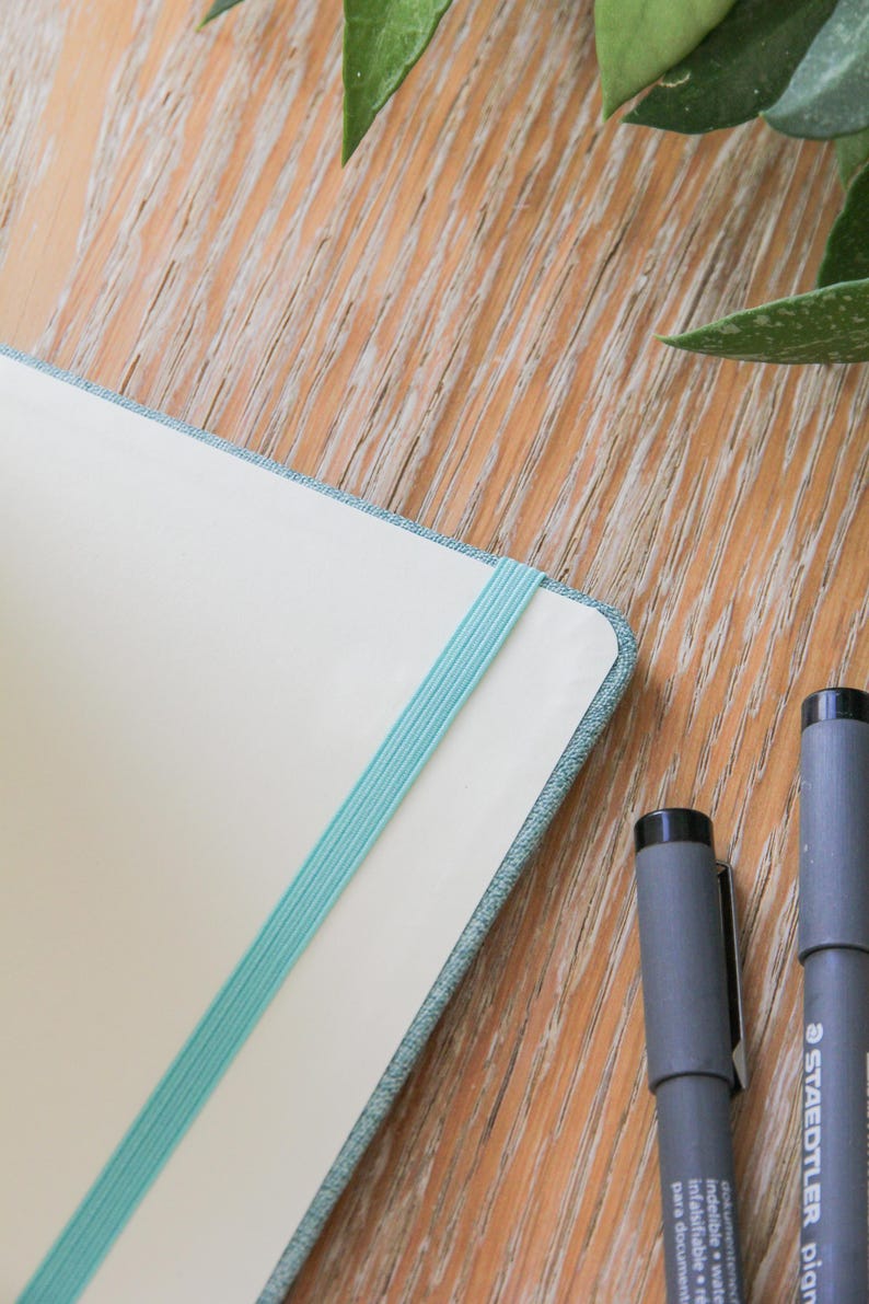 Puede incluir: Un cuaderno con una banda el&aacute;stica azul claro y una cubierta azul claro est&aacute; abierto sobre una superficie de madera. Dos rotuladores grises Staedtler est&aacute;n junto al cuaderno. Hojas verdes en el fondo.