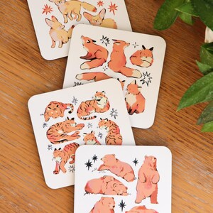 May include: Set of four square coasters with animal illustrations. Each coaster features a different animal: foxes, tigers, bears, and rabbits. The animals are in shades of orange and brown, with star accents on a white background.