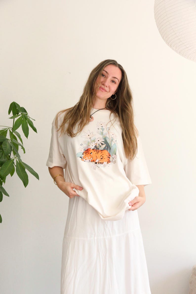 May include: Cream-colored t-shirt featuring a watercolor illustration of a sleeping orange tiger surrounded by white flowers and green leaves. The shirt is paired with a long, white skirt. The model is smiling.