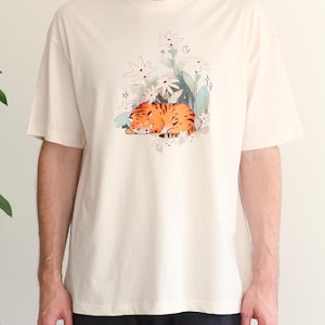May include: Cream-colored t-shirt featuring a watercolor illustration of a sleeping orange tiger among white flowers and green foliage. The short-sleeved shirt has a crew neck and a relaxed fit, suitable for casual wear.