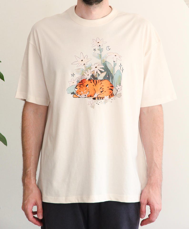 May include: Cream-colored t-shirt featuring a watercolor illustration of a sleeping orange tiger among white flowers and green foliage. The short-sleeved shirt has a crew neck and a relaxed fit, suitable for casual wear.
