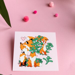 May include: A pink card featuring a charming illustration of five orange foxes surrounded by green plants and pink flowers. The foxes are in various poses, some looking up, some looking down, and some looking at each other. A pink heart is in the top left corner of the card.