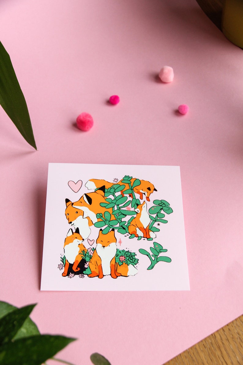 May include: A pink card featuring a charming illustration of five orange foxes surrounded by green plants and pink flowers. The foxes are in various poses, some looking up, some looking down, and some looking at each other. A pink heart is in the top left corner of the card.