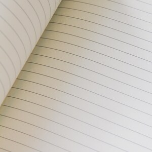 Puede incluir: Cuaderno abierto con p&aacute;ginas rayadas. Las p&aacute;ginas son de color blanquecino con finas l&iacute;neas horizontales grises. El cuaderno est&aacute; abierto, revelando las p&aacute;ginas interiores y la curvatura de la encuadernaci&oacute;n.