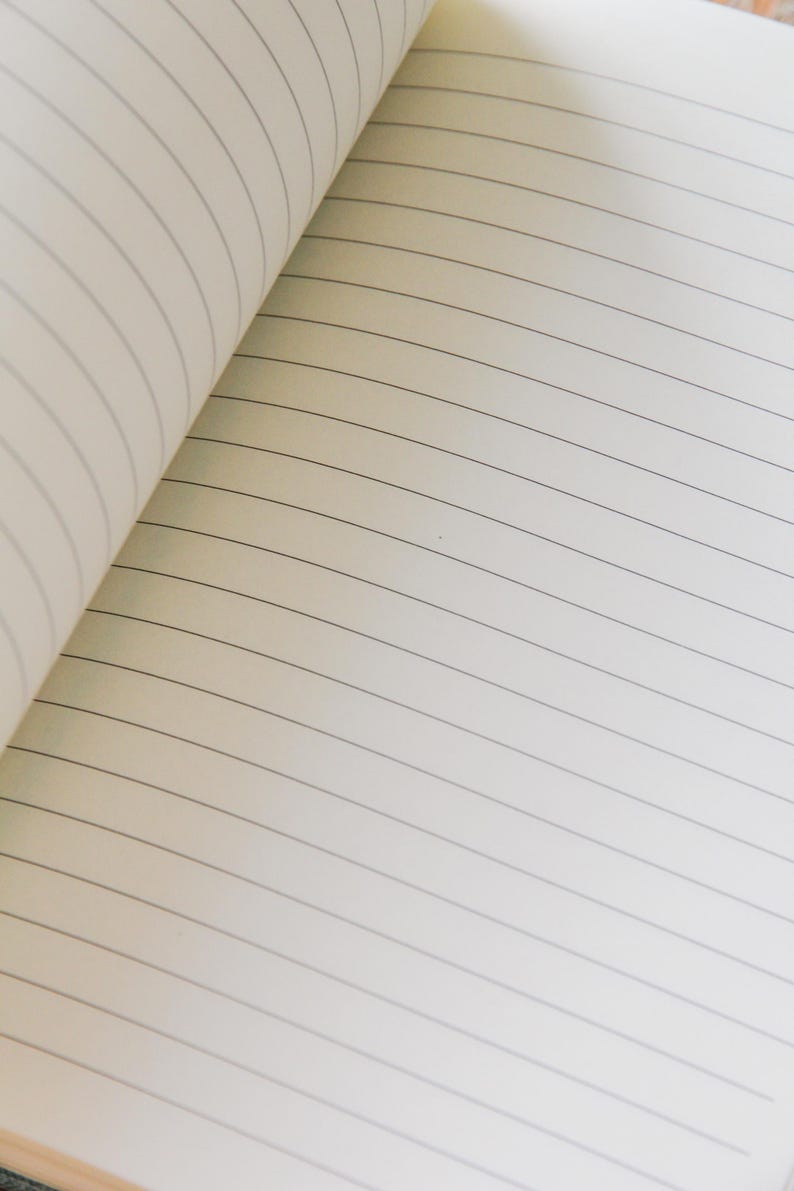 Puede incluir: Cuaderno abierto con p&aacute;ginas rayadas. Las p&aacute;ginas son de color blanquecino con finas l&iacute;neas horizontales grises. El cuaderno est&aacute; abierto, revelando las p&aacute;ginas interiores y la curvatura de la encuadernaci&oacute;n.