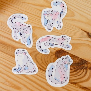 Puede incluir: Cuatro pegatinas coloridas con ilustraciones de leopardos de las nieves en varias poses. Las pegatinas est&aacute;n sobre una superficie de madera clara.