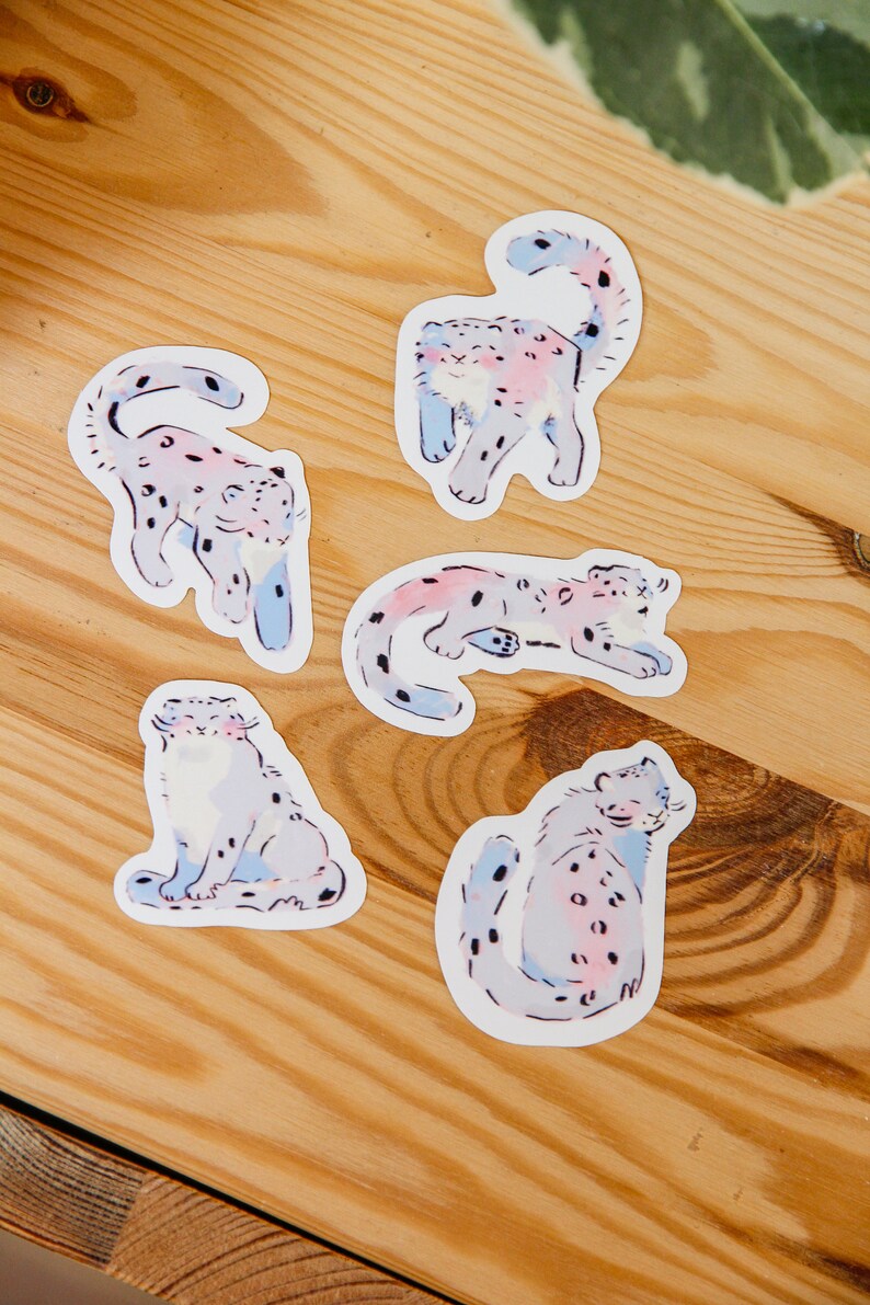 Puede incluir: Cuatro pegatinas coloridas con ilustraciones de leopardos de las nieves en varias poses. Las pegatinas est&aacute;n sobre una superficie de madera clara.
