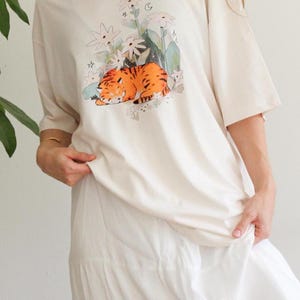 May include: Cream-colored t-shirt featuring a graphic of a sleeping orange tiger amidst white flowers and green foliage. The shirt has short sleeves and a relaxed fit, suitable for casual wear. The overall aesthetic is soft and whimsical.