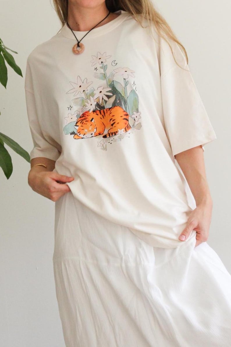 May include: Cream-colored t-shirt featuring a graphic of a sleeping orange tiger amidst white flowers and green foliage. The shirt has short sleeves and a relaxed fit, suitable for casual wear. The overall aesthetic is soft and whimsical.