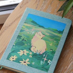Puede incluir: Un cuaderno azul claro con una ilustraci&oacute;n de un gato en la portada. La ilustraci&oacute;n representa un gato color crema sentado en un campo de flores bajo un cielo azul. El cuaderno est&aacute; sobre una superficie de madera.