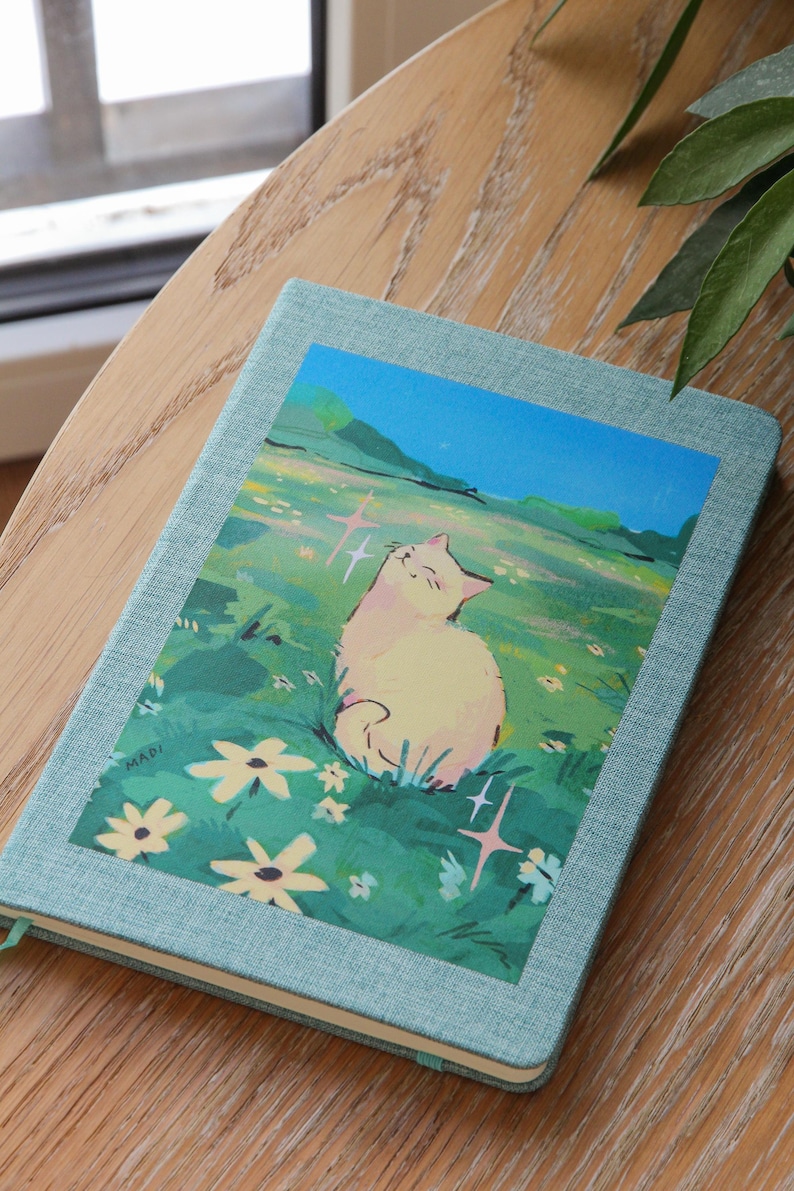 Puede incluir: Un cuaderno azul claro con una ilustraci&oacute;n de un gato en la portada. La ilustraci&oacute;n representa un gato color crema sentado en un campo de flores bajo un cielo azul. El cuaderno est&aacute; sobre una superficie de madera.