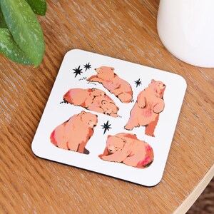 May include: White square coaster featuring illustrations of pink bears in various poses, including standing, sleeping, and sitting. Black star accents are scattered around the bears. The coaster is on a wooden surface.