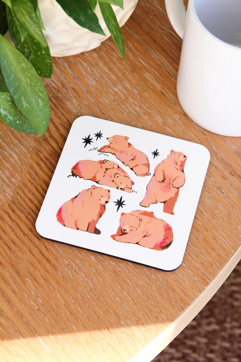 May include: White square coaster featuring illustrations of pink bears in various poses, including standing, sleeping, and sitting. Black star accents are scattered around the bears. The coaster is on a wooden surface.
