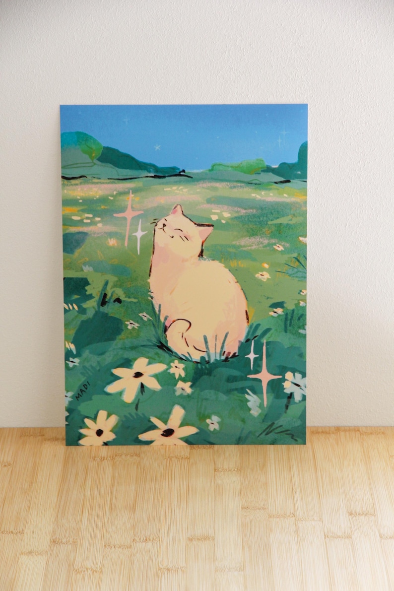 May include: A whimsical art print featuring a cream-colored cat basking in a field of flowers under a starry night sky. The illustration has a soft, dreamy aesthetic with pastel colors and sparkling accents. The artist's signature, MADI, is visible.