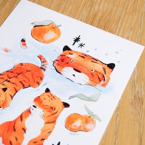 May include: A white card featuring three orange tigers, two oranges, and two black stars. The tigers are depicted in a cartoon style. The card is signed "MADI".