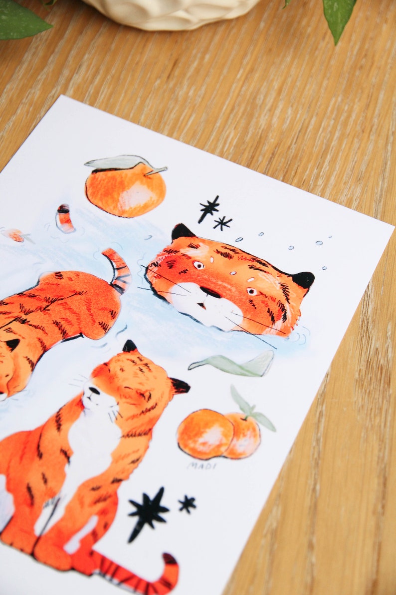 May include: A white card featuring three orange tigers, two oranges, and two black stars. The tigers are depicted in a cartoon style. The card is signed "MADI".