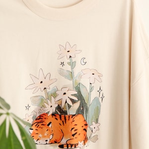 May include: Cream-colored t-shirt featuring a whimsical illustration of a sleeping tiger amidst stylized flowers, stars, and a crescent moon. The tiger is orange with black stripes. The shirt has a relaxed fit and a crew neck.