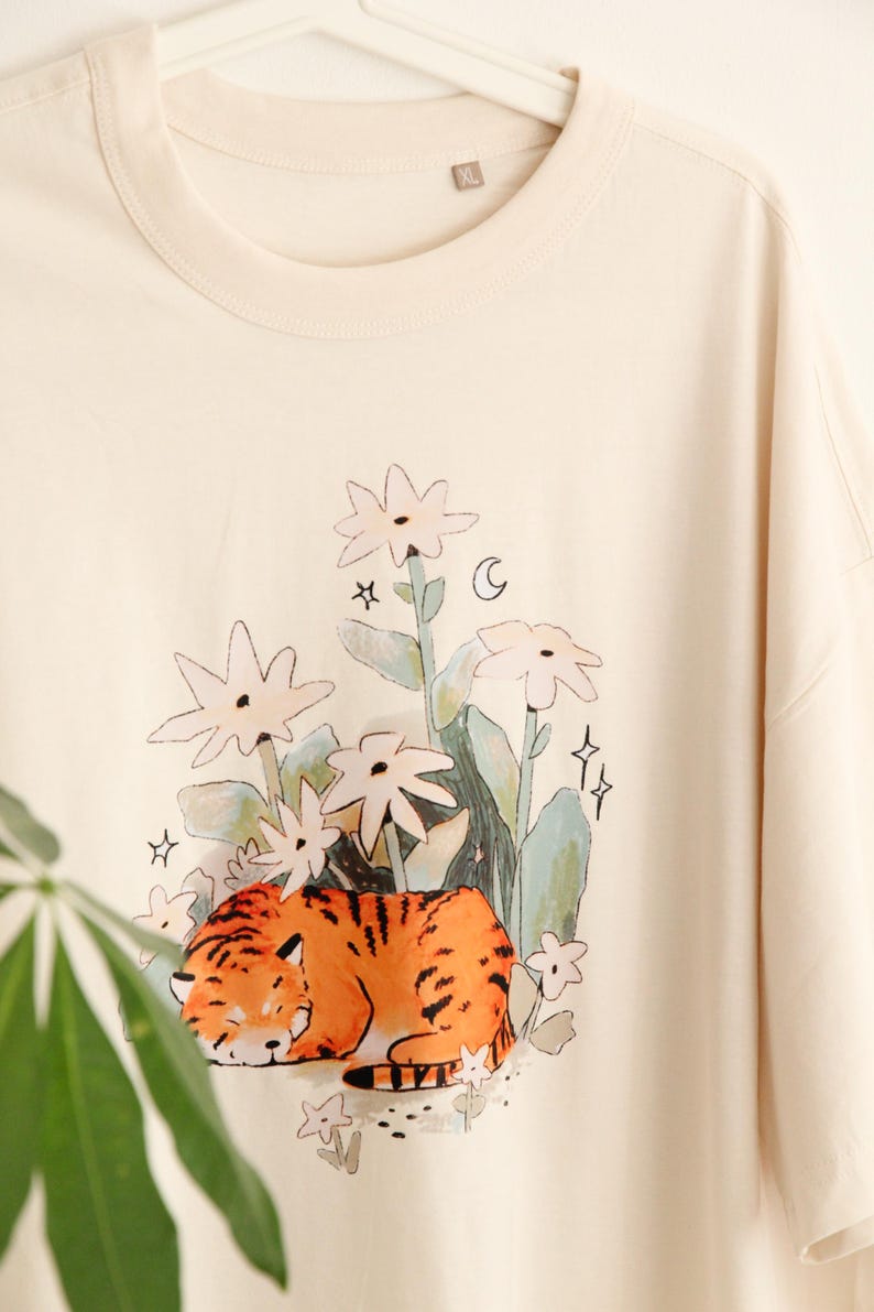 May include: Cream-colored t-shirt featuring a whimsical illustration of a sleeping tiger amidst stylized flowers, stars, and a crescent moon. The tiger is orange with black stripes. The shirt has a relaxed fit and a crew neck.