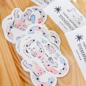 Puede incluir: Un conjunto de 6 pegatinas de leopardo de las nieves de estilo acuarela en una bolsa de pl&aacute;stico transparente. Las pegatinas son de color rosa, azul y blanco con manchas negras. La bolsa tiene una etiqueta que dice "Snow Leopard Stickerpack madivoid".