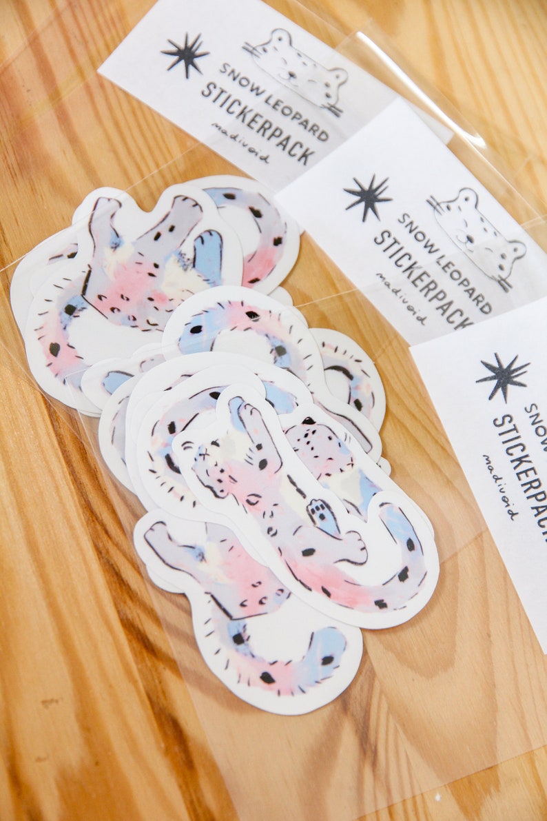 Puede incluir: Un conjunto de 6 pegatinas de leopardo de las nieves de estilo acuarela en una bolsa de pl&aacute;stico transparente. Las pegatinas son de color rosa, azul y blanco con manchas negras. La bolsa tiene una etiqueta que dice "Snow Leopard Stickerpack madivoid".