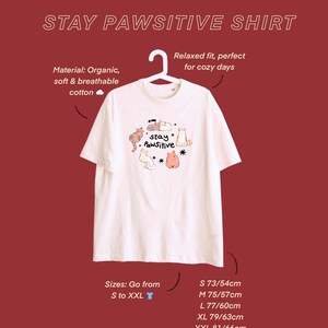 Puede incluir: Camiseta blanca con el texto "STAY PAWSITIVE SHIRT" y un gr&aacute;fico de gatos de dibujos animados. Fabricada en algod&oacute;n org&aacute;nico, suave y transpirable. Tallas de la S a la XXL, con medidas en cm: S 73/54cm, M 75/57cm, L 77/60cm, XL 79/63cm, XXL 81/66cm.
