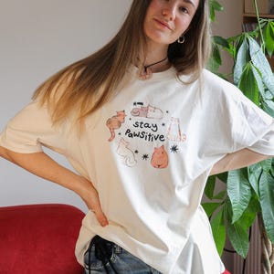 Puede incluir: Camiseta color crema con una ilustraci&oacute;n de gato y el texto "stay Pawsitive". El dise&ntilde;o incluye varios gatos de dibujos animados en diferentes poses y colores. La camiseta es de estilo casual y oversize.