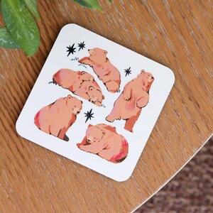 May include: White square coaster featuring illustrations of brown bears in various poses. The bears are depicted with pink accents and are surrounded by black star and wavy line designs. A charming and whimsical design.
