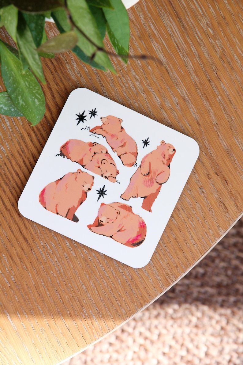 May include: White square coaster featuring illustrations of brown bears in various poses. The bears are depicted with pink accents and are surrounded by black star and wavy line designs. A charming and whimsical design.