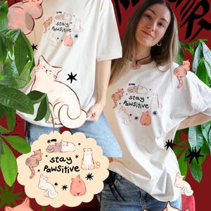 Puede incluir: Camiseta de color crema con una ilustraci&oacute;n de gato y las palabras "stay Pawsitive". La camiseta presenta varios dise&ntilde;os de gatos de dibujos animados en tonos de rosa, marr&oacute;n y blanco. El fondo es rojo oscuro con follaje verde.