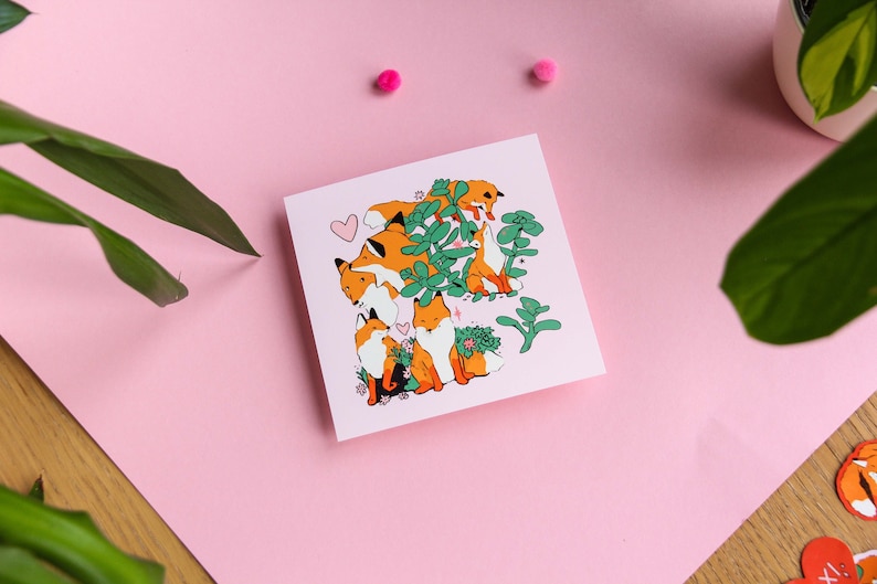May include: A pink card featuring a charming illustration of foxes nestled in a green bush. The foxes are orange with white markings and there are small pink hearts dotted around them.