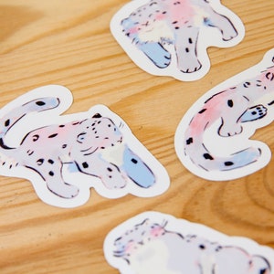 Puede incluir: Cinco pegatinas coloridas con ilustraciones de leopardos de las nieves. Las pegatinas est&aacute;n sobre una superficie de madera marr&oacute;n claro. Los leopardos de las nieves est&aacute;n dibujados en un estilo caprichoso con lavados de acuarela rosa, azul y blanco.