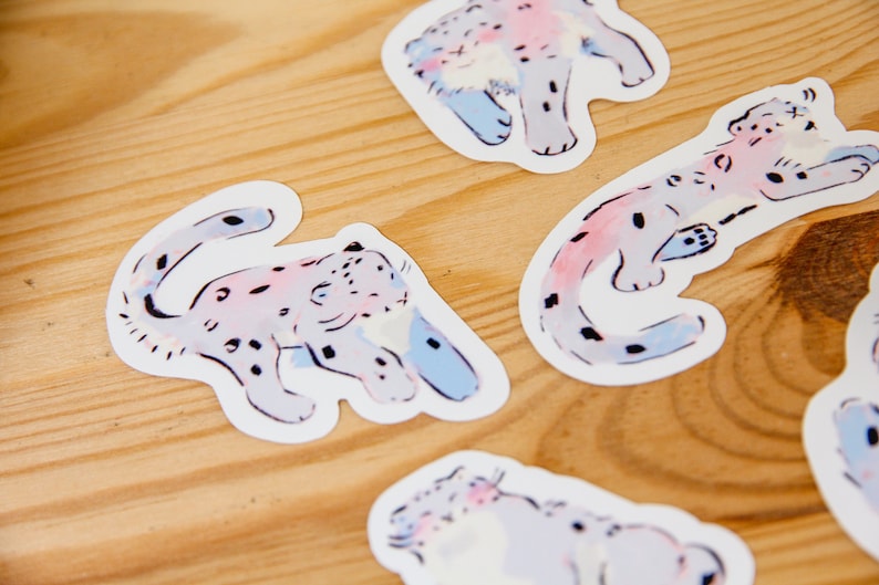 Puede incluir: Cinco pegatinas coloridas con ilustraciones de leopardos de las nieves. Las pegatinas est&aacute;n sobre una superficie de madera marr&oacute;n claro. Los leopardos de las nieves est&aacute;n dibujados en un estilo caprichoso con lavados de acuarela rosa, azul y blanco.