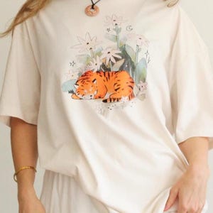 May include: Cream-colored t-shirt featuring a sleeping tiger illustration in orange and black stripes, surrounded by white flowers and green foliage. The shirt has short sleeves and a relaxed fit, suitable for casual wear.