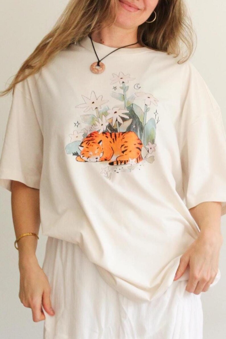 May include: Cream-colored t-shirt featuring a sleeping tiger illustration in orange and black stripes, surrounded by white flowers and green foliage. The shirt has short sleeves and a relaxed fit, suitable for casual wear.