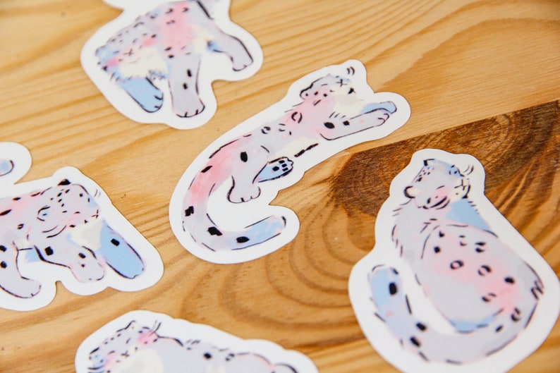 Puede incluir: Cinco pegatinas coloridas con ilustraciones de leopardos de las nieves en varias poses. Las pegatinas est&aacute;n sobre un fondo de madera clara.