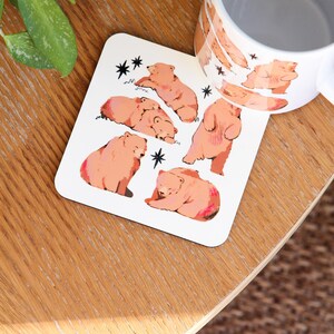 May include: White coaster with illustrations of brown bears in various poses, with black star accents. A matching mug with bear illustrations is also visible. The items are on a wooden surface.