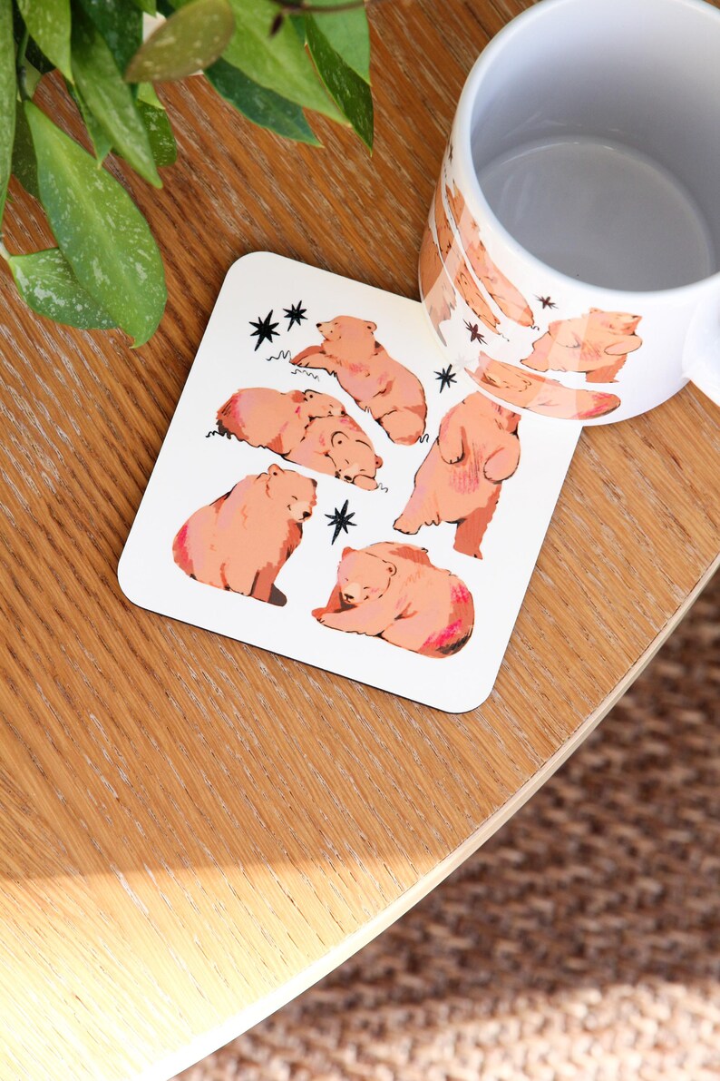 May include: White coaster with illustrations of brown bears in various poses, with black star accents. A matching mug with bear illustrations is also visible. The items are on a wooden surface.