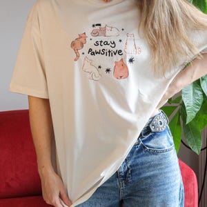 Puede incluir: Camiseta beige con un gr&aacute;fico de gatos. El dise&ntilde;o presenta varios gatos de dibujos animados en varias poses, rodeados de estrellas, y las palabras "stay Pawsitive". La camiseta tiene mangas cortas y cuello redondo.