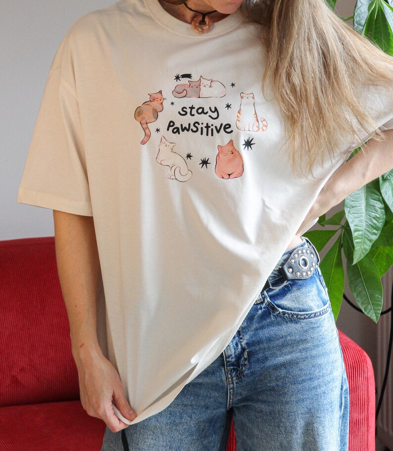 Puede incluir: Camiseta beige con un gr&aacute;fico de gatos. El dise&ntilde;o presenta varios gatos de dibujos animados en varias poses, rodeados de estrellas, y las palabras "stay Pawsitive". La camiseta tiene mangas cortas y cuello redondo.