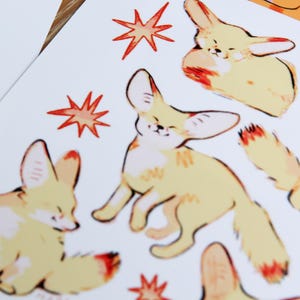 May include: A sheet of stickers featuring illustrations of fennec foxes in various poses. The foxes are depicted in a light yellow color with orange accents. The background is white with red starburst designs.