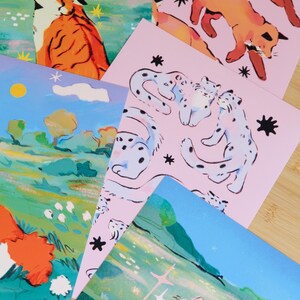 May include: A collection of art prints featuring illustrations of animals. The prints showcase a tiger, foxes, snow leopards, and a cat, with a color palette of orange, pink, blue, and green. The artwork has a whimsical, illustrative style.