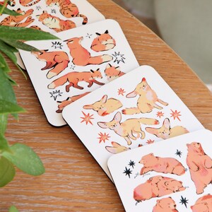 May include: Set of four square coasters featuring illustrations of various animals. The coasters showcase orange tigers, foxes, fennec foxes, and bears, with star and lightning bolt accents. The coasters are on a wooden surface.